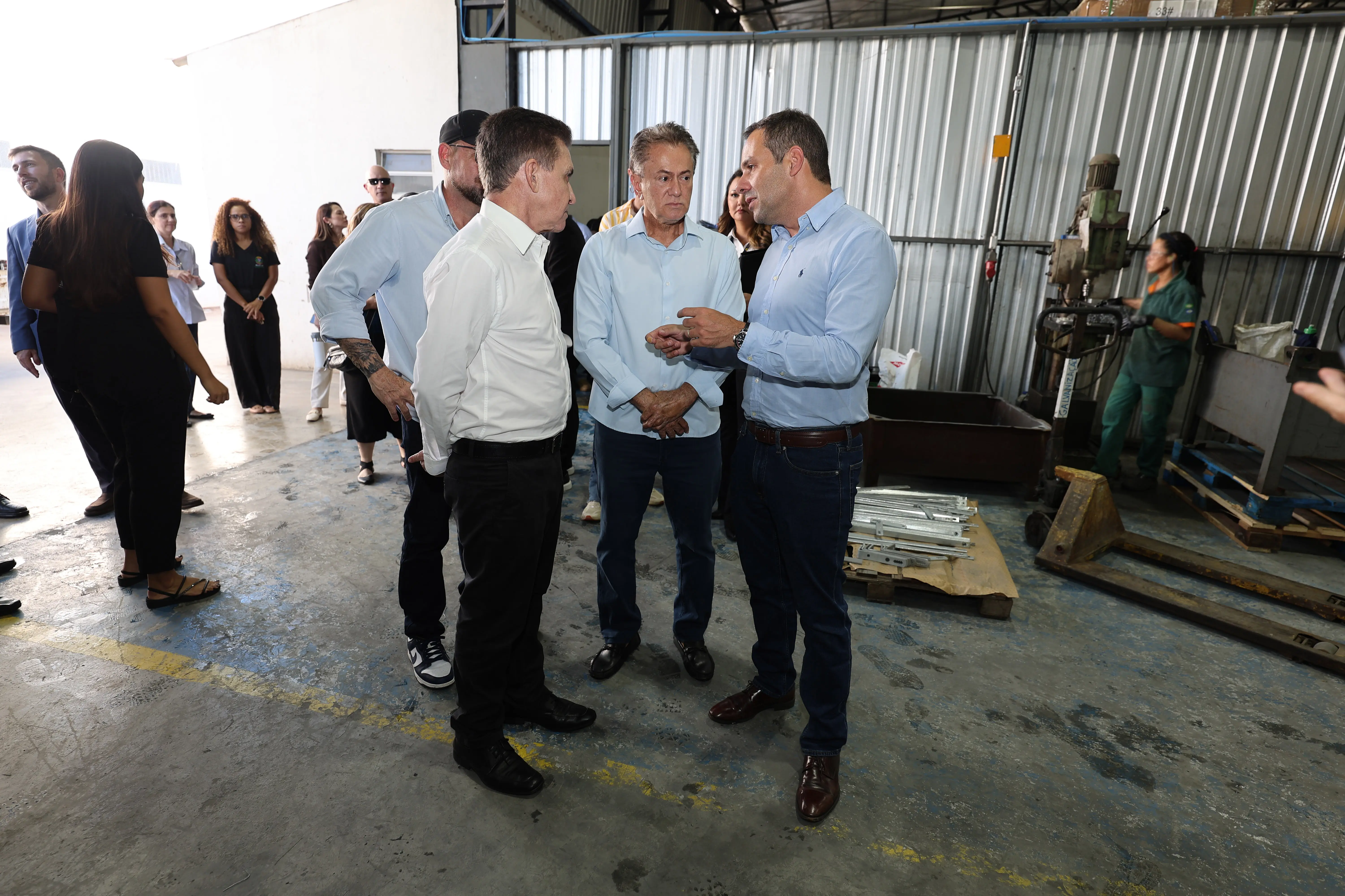Três homens de camisa social conversam em galpão industrial. Ao fundo, funcionária de macacão verde opera equipamento. Grupo de pessoas aguarda ao lado.