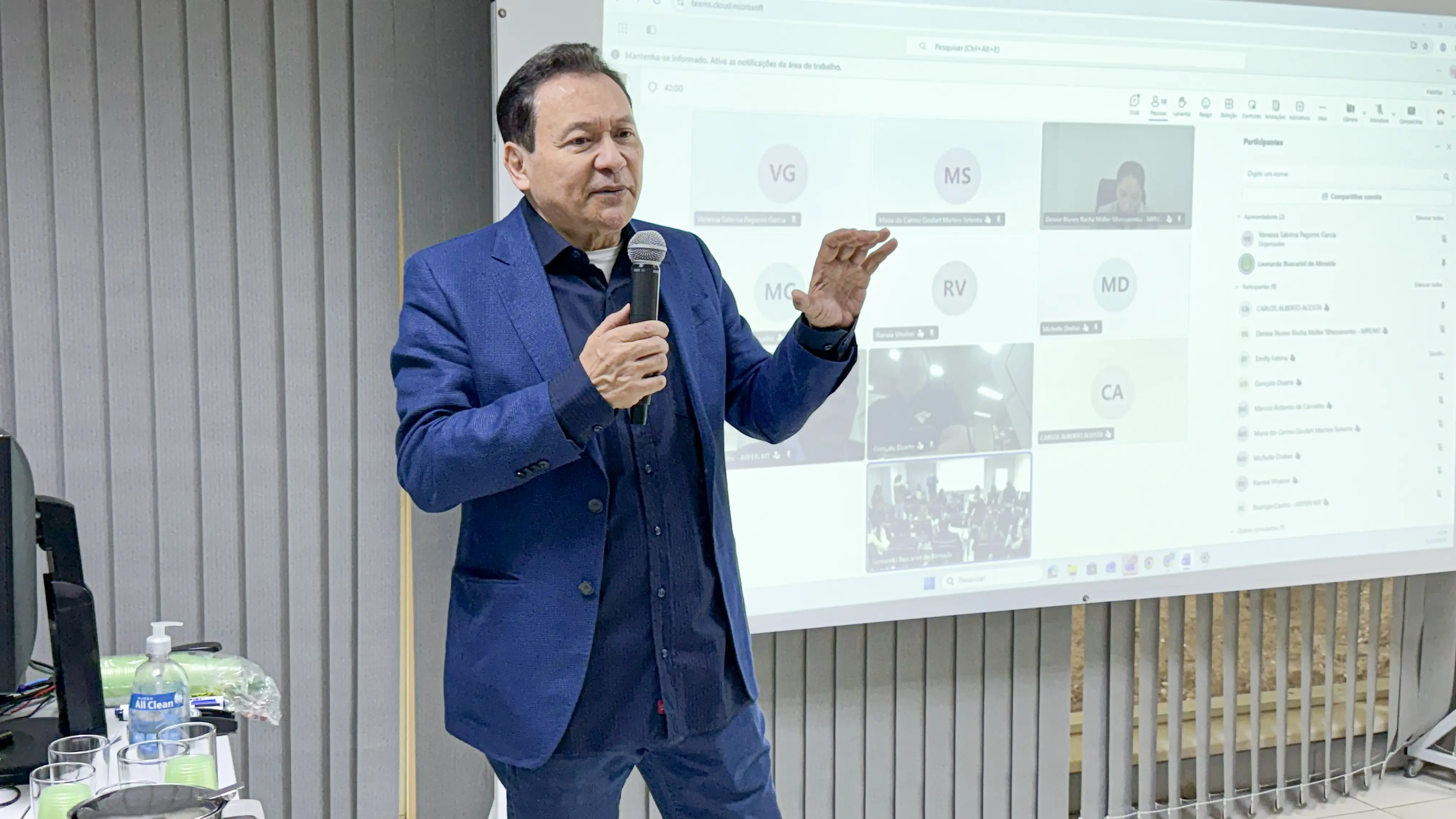 Homem de cabelos escuros, veste terno azul, fala ao microfone. Ao fundo, tela exibe videoconferência com vários participantes.