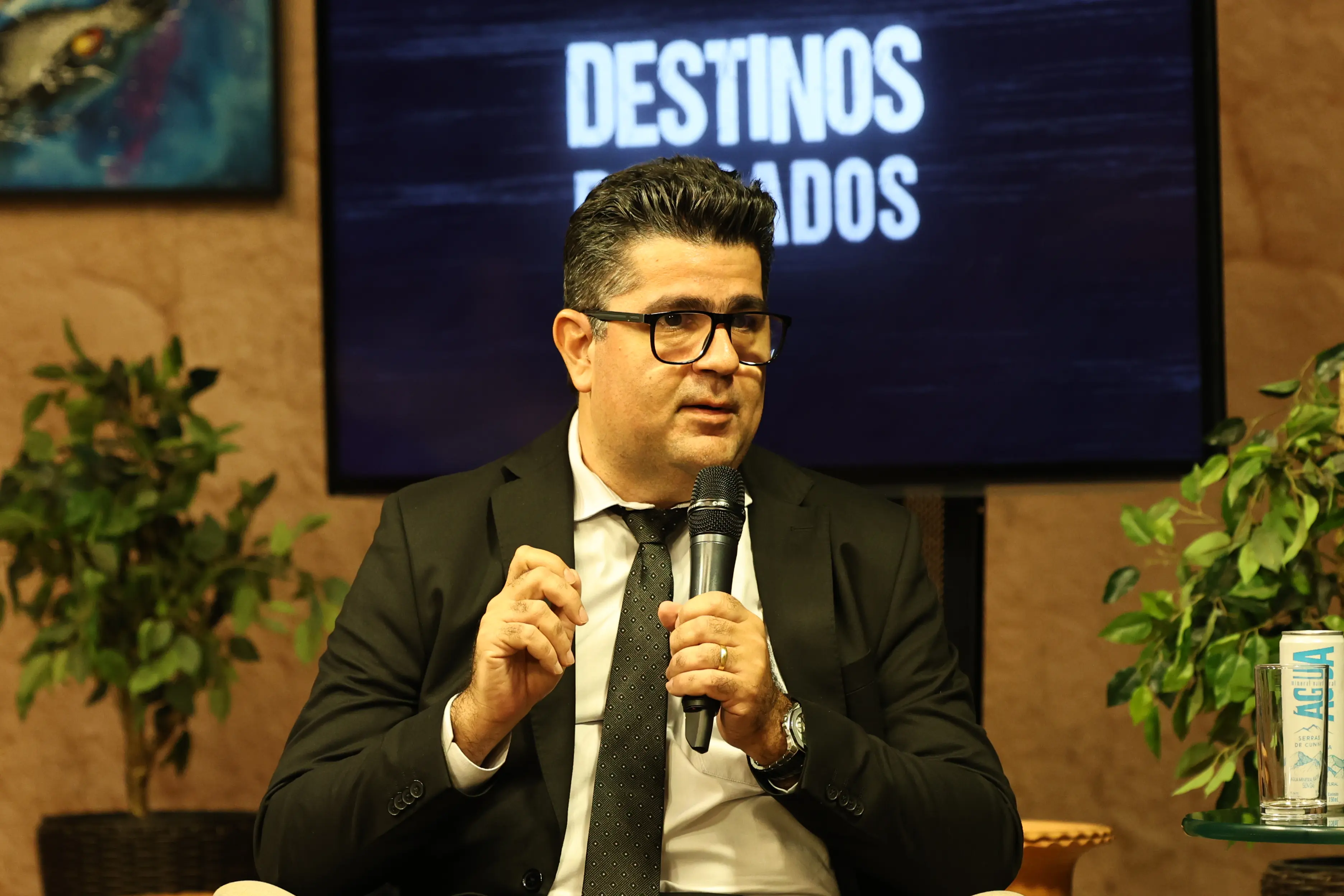 A imagem mostra o juiz Marcos Terêncio durante sua participação no debate sobre violência doméstica. Ele é um homem de pele clara, cabelos grisalhos nas temporas, olhos escuros e usa óculos de grau. Está segurando o microfone com a mão direita. Veste terno e gravata pretos e camisa branca. 