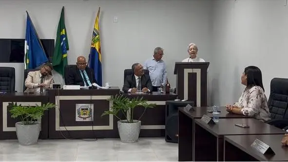 Visão ampla do plenário. Uma mulher de cabelos brancos discursa no púlpito. Outras pessoas estão sentadas e em pé atrás da bancada principal, que exibe um brasão e plantas nas laterais.