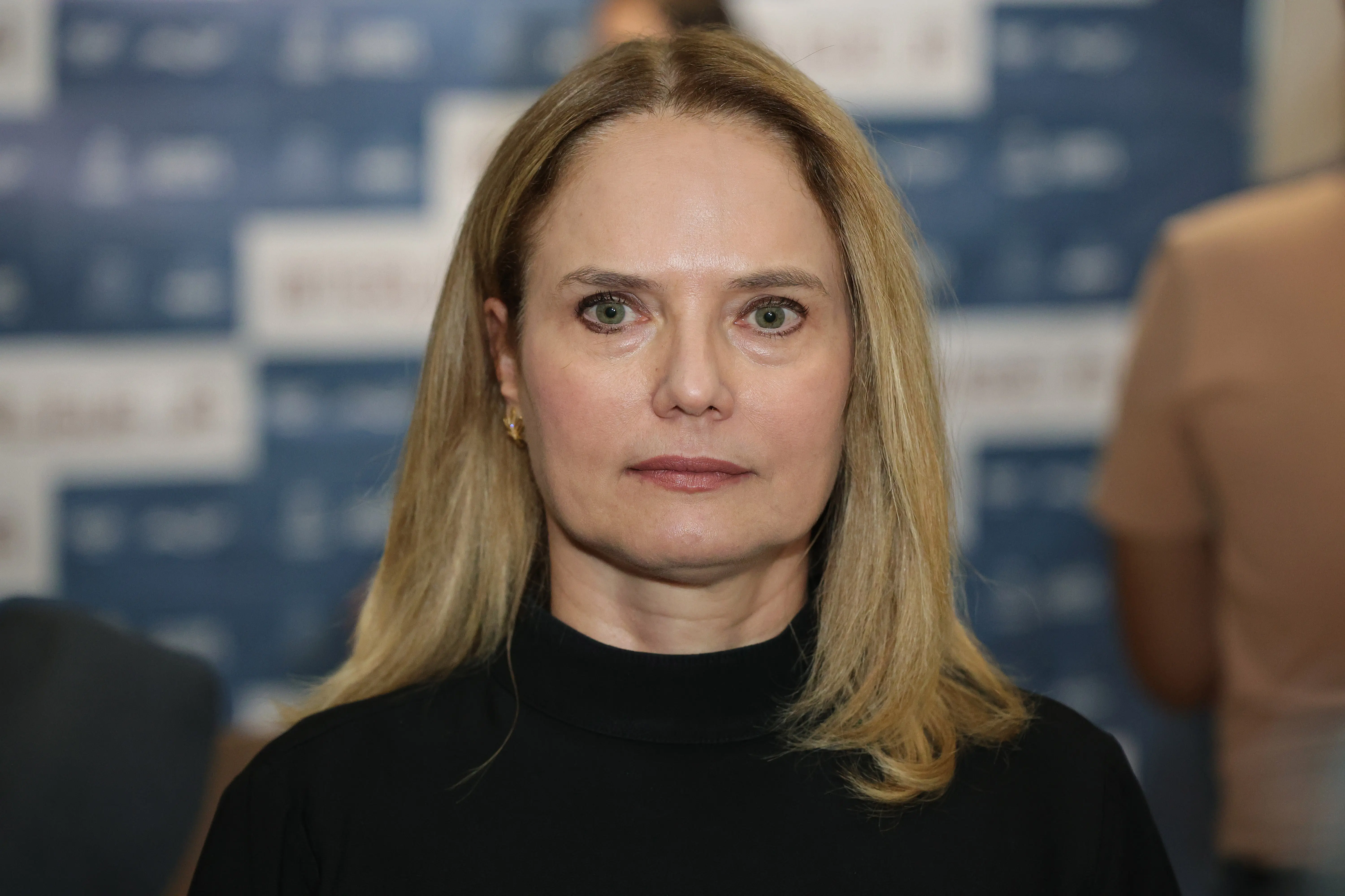 Mulher de cabelos loiros lisos, blusa preta de gola alta, olha para a câmera. Ao fundo, painel institucional azul e branco.