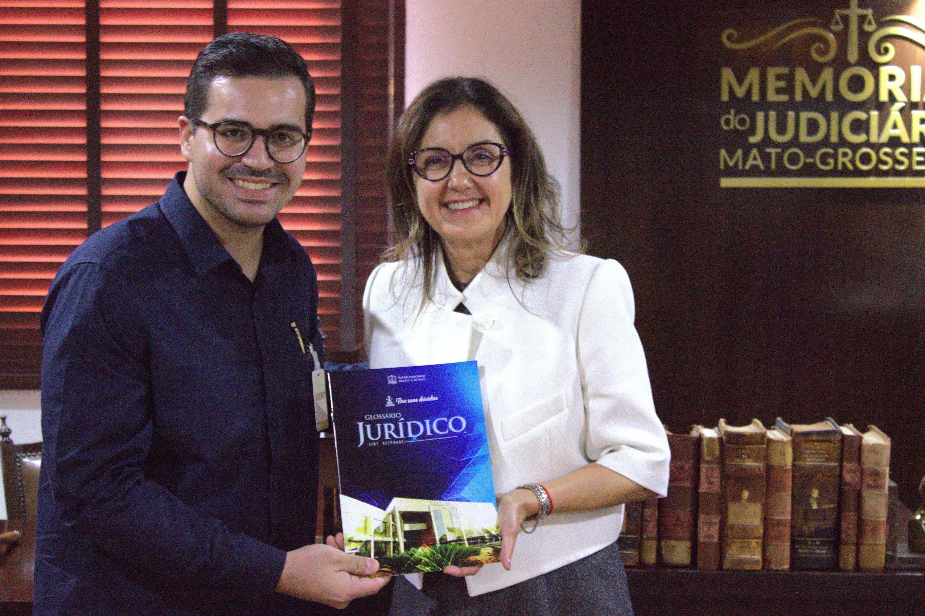 Homem de óculos redondos, camisa preta, e mulher de blazer branco seguram um Glossário Jurídico. Ao fundo, painel do Memorial do Judiciário Mato-Grossense.