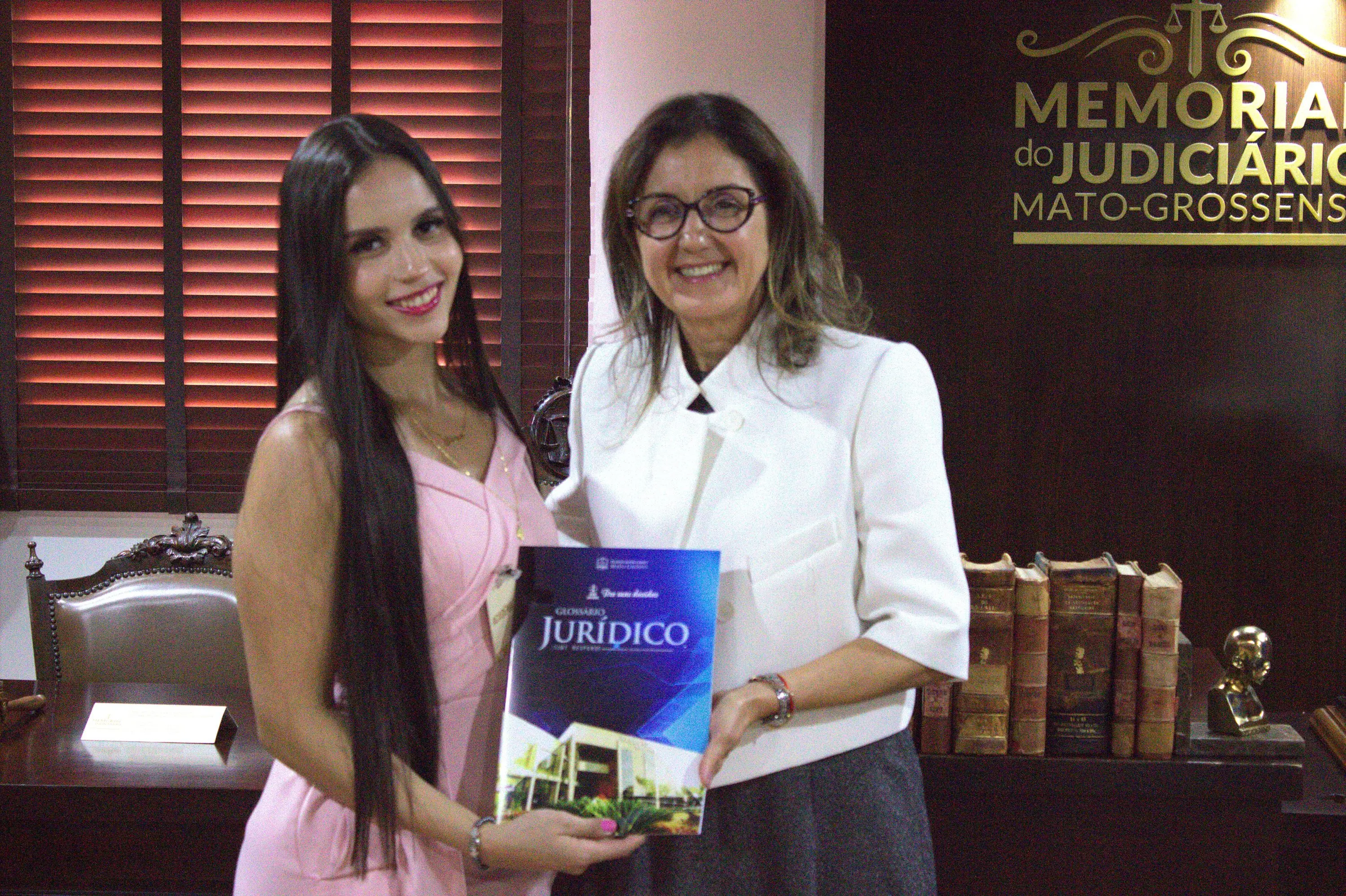 Jovem de cabelos longos escuros, vestido rosa, e mulher de óculos, blazer branco, seguram juntas um Glossário Jurídico. Ao fundo, painel do Memorial do Judiciário Mato-Grossense.