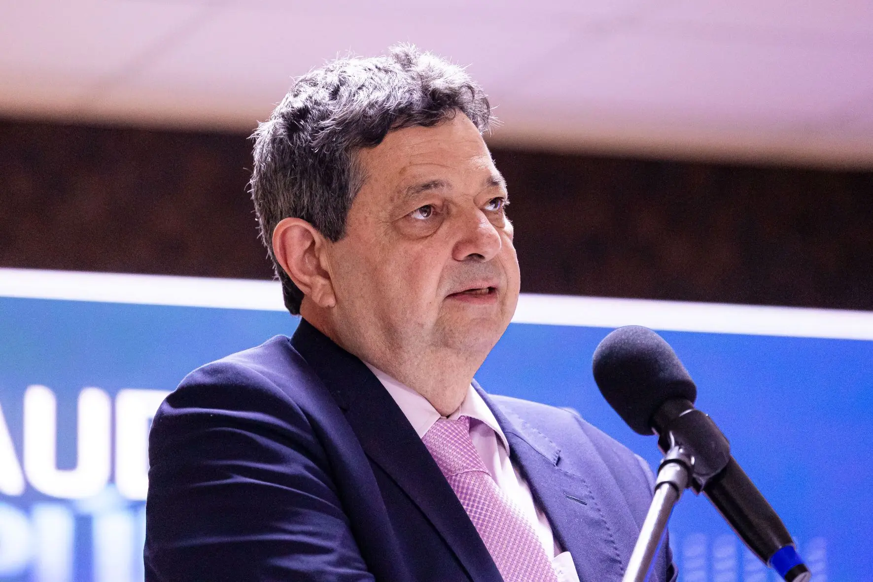 Foto horizontal que mostra o desembargador José Luiz Leite Lindote do busto pra cima, falando ao microfone. Ele é um homem branco, de cabelos grisalhos, usando camisa branca, gravata rosa e terno azul marinho.