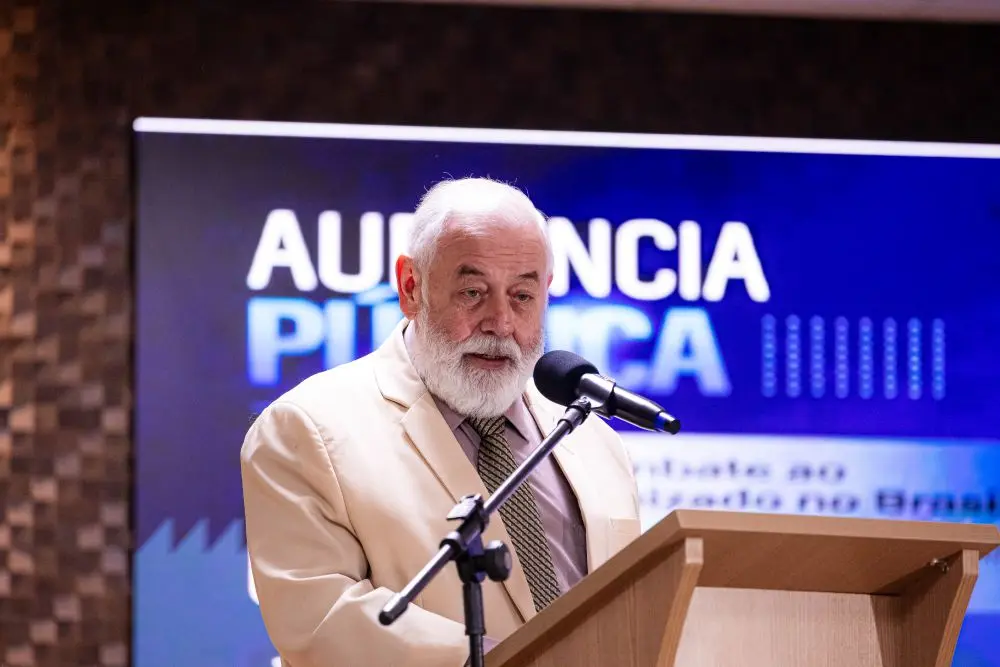 Foto horizontal que mostra o presidente do TJMT, desembargador José Zuquim, falando ao microfone. Ele é um senhor de pele branca, cabelos e barba brancos, usando terno cor creme e gravata marrom. Ao fundo, um telão com as informações da audiência pública.