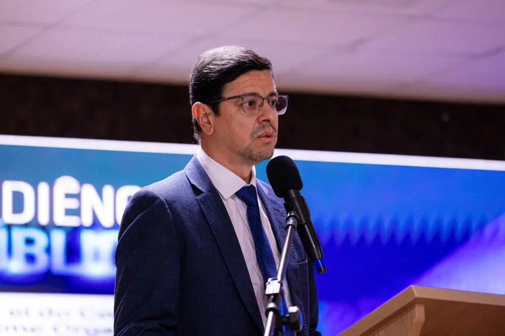 Foto horizontal que mostra o promotor de justiça Renee do Ó do busto pra cima, falando ao microfone. Ele é um homem branco, de cabelo preto, usando camisa branca, gravata e terno azul marinho e óculos de grau. Atrás dele é possível ver parte de um telão.