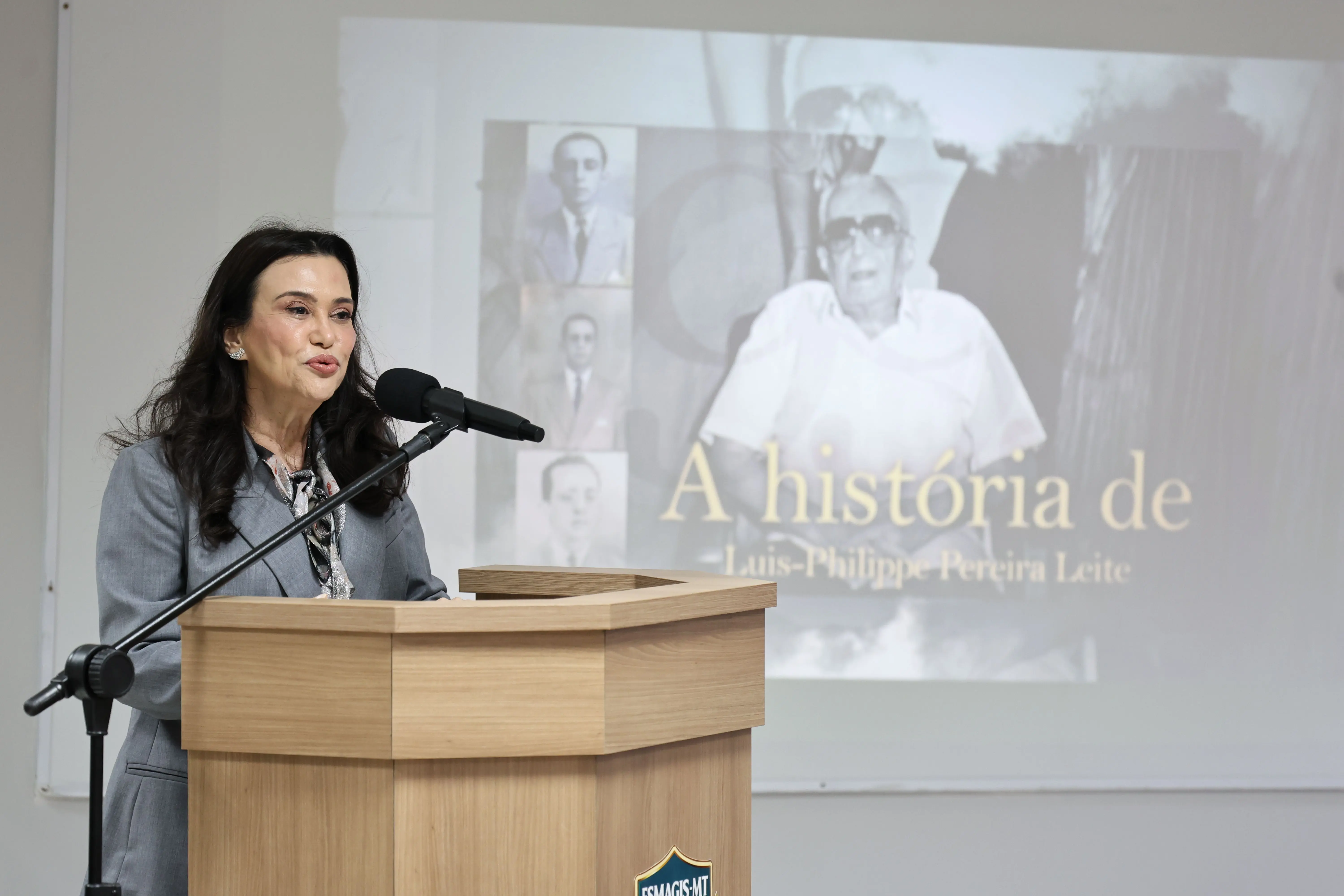 Mulher de longos cabelos pretos fala ao microfone em um púlpito de madeira clara, vestindo um blazer cinza. Ao fundo, uma tela de projeção exibe imagens antigas em preto e branco. No canto inferior do púlpito, há um pequeno emblema azul e dourado com a inscrição parcial 