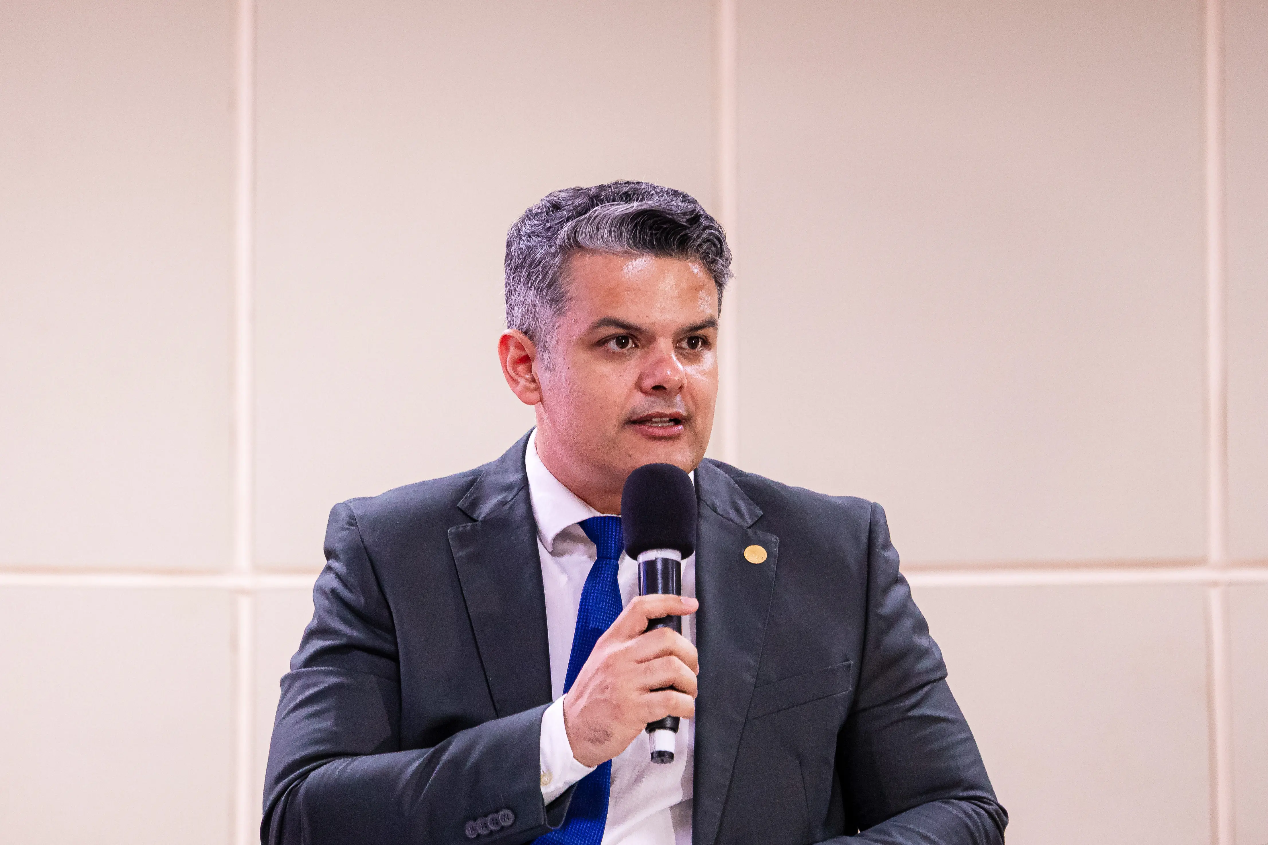 Homem de terno escuro, camisa branca e gravata azul, com cabelos grisalhos curtos, segura um microfone e fala. Ele tem um pin dourado na lapela. O fundo é uma parede de painéis claros.
