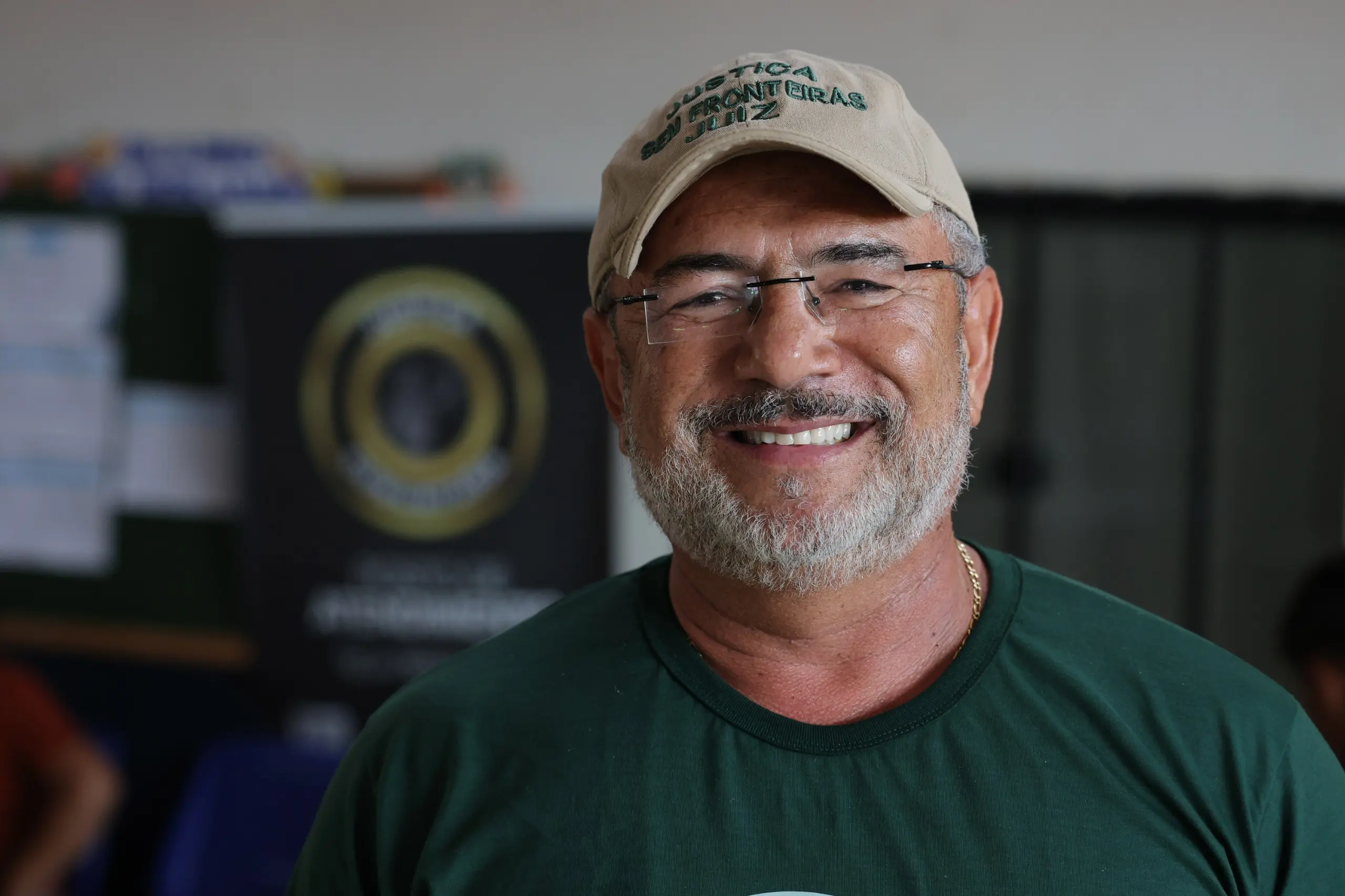 Homem usando boné, óculos e camiseta da Expedição Justiça Sem Fronteiras posa para fotografia em ambiente interno.