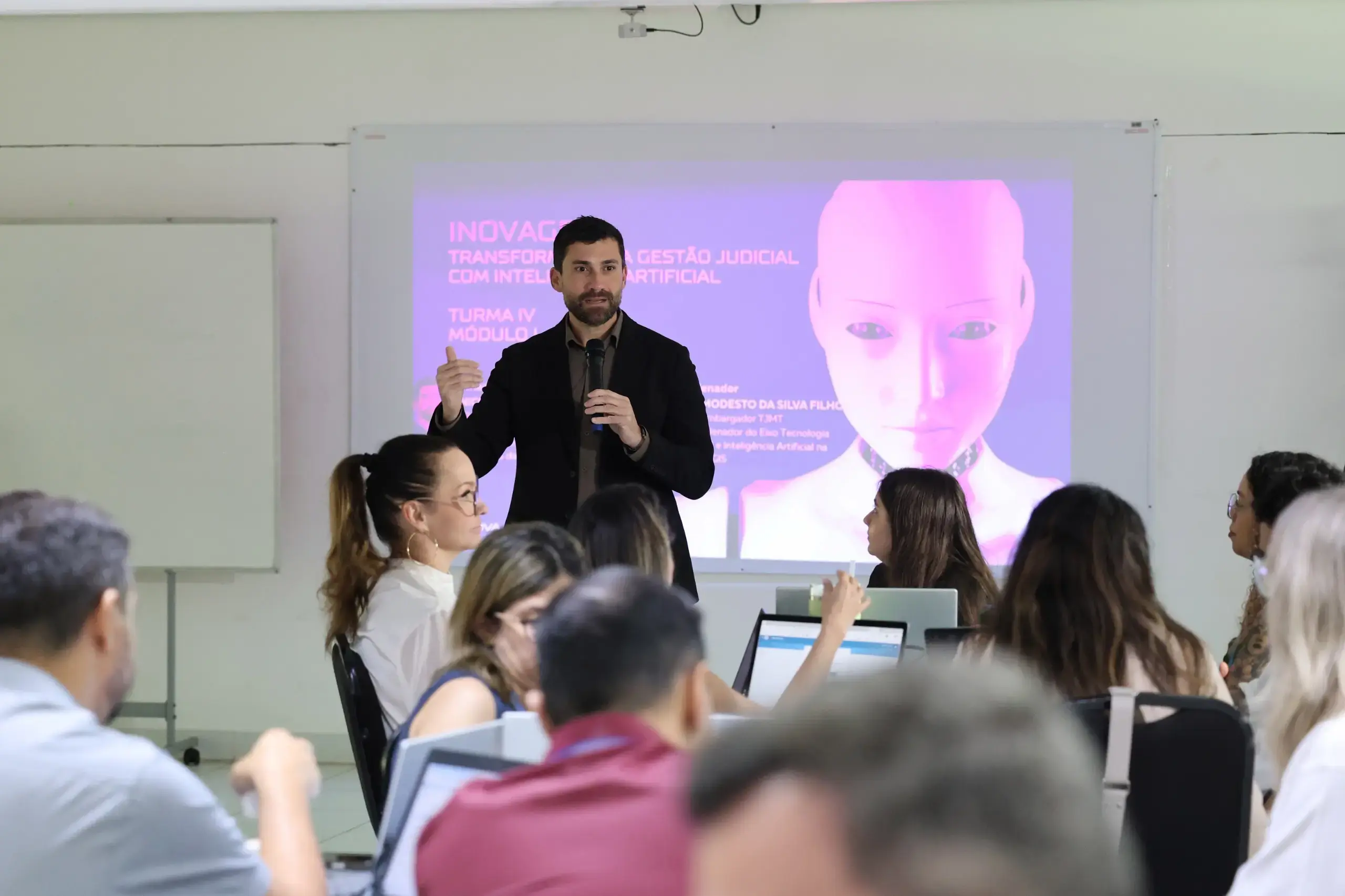 Homem de paletó escuro fala ao microfone diante de uma plateia. Ao fundo, uma tela de projeção exibe um slide lilás com o rosto de um humanoide e o texto 