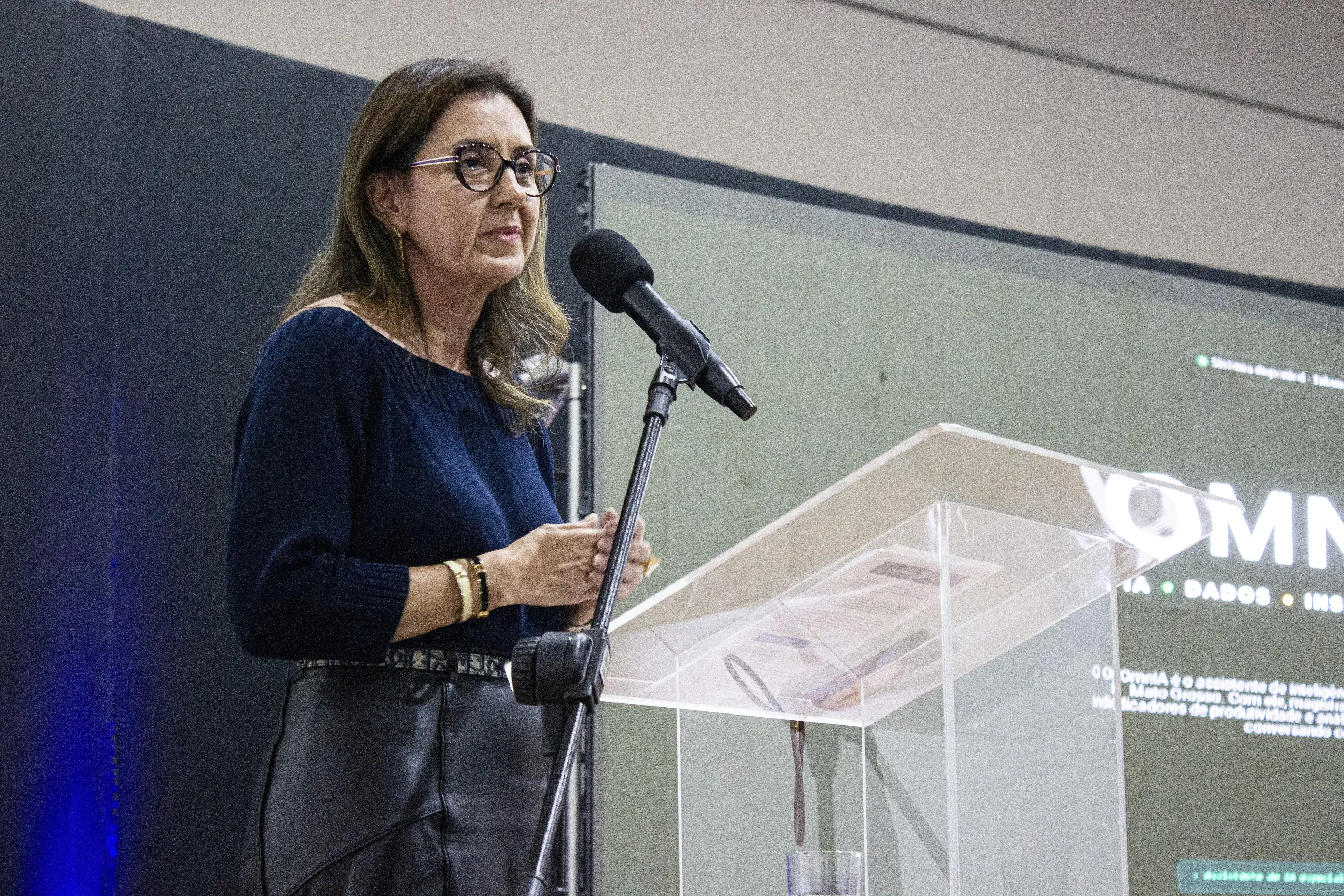 Mulher de óculos e blusa azul-escura discursa em um púlpito de acrílico transparente com microfone à frente. Ao fundo, uma tela projeta um slide esverdeado com a palavra parcial 