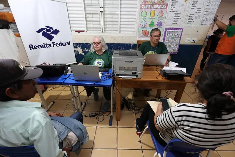 Dois atendentes com notebooks diante de um banner da Receita Federal, prestando atendimento a um homem e uma mulher sentados.
