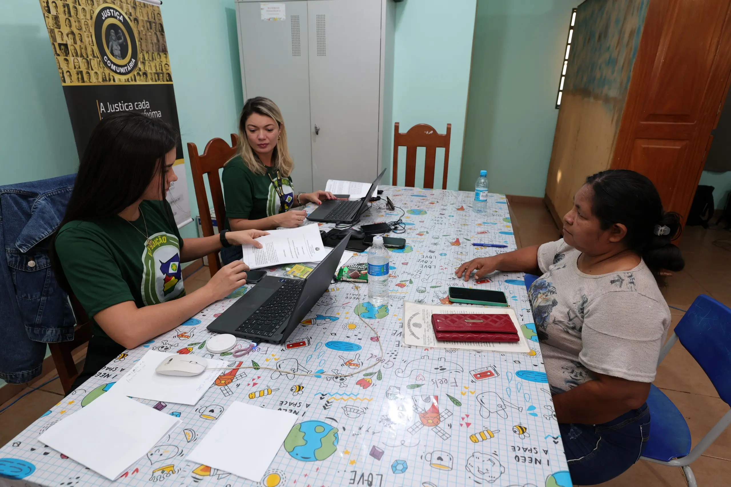 A imagem mostra duas atendentes de camiseta verde com notebooks em uma mesa estampada, prestando atendimento a uma mulher sentada à frente delas.
