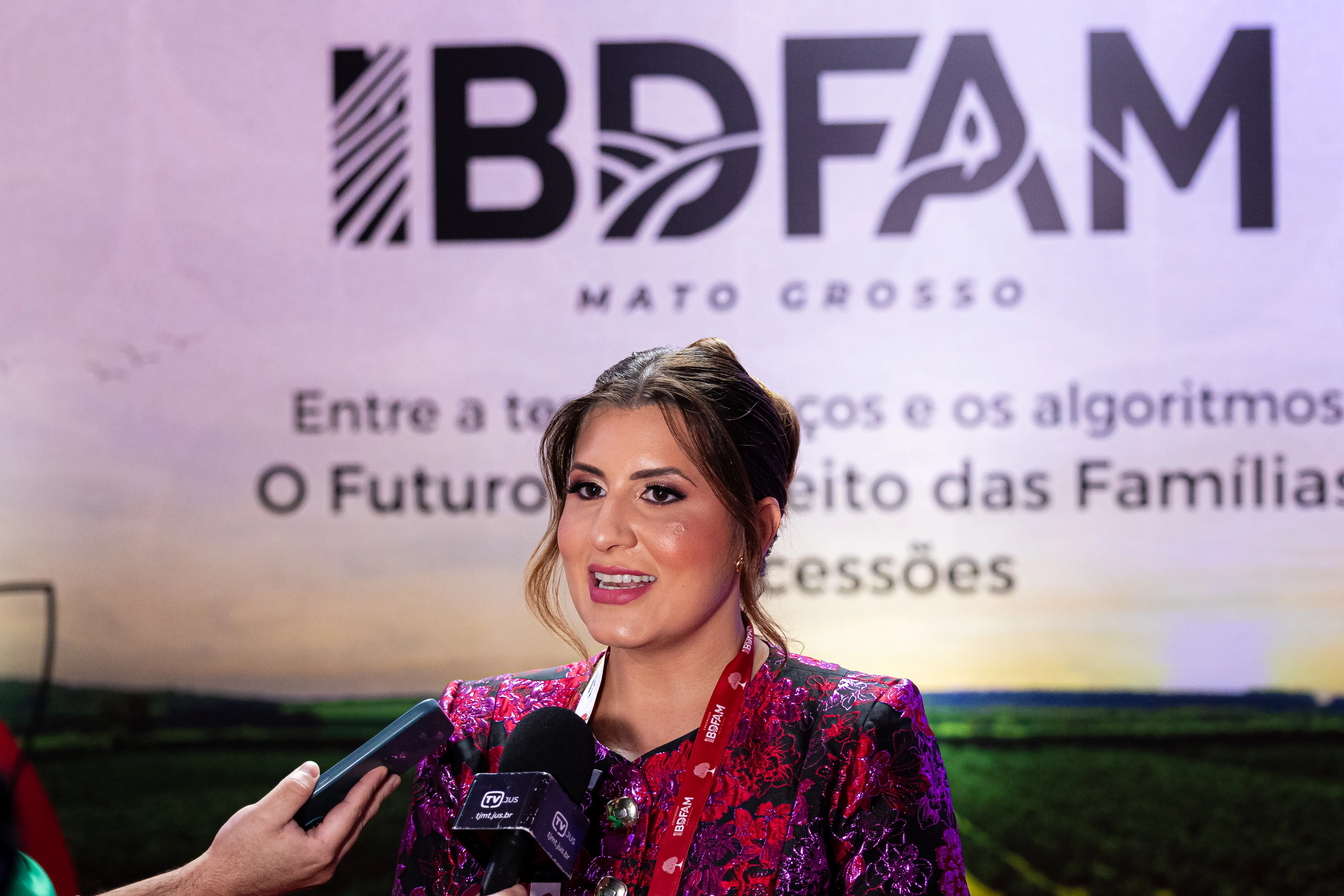 Mulher de cabelo preso e blazer floral brilhante concede entrevista, falando ao microfone da TV Jus. Ao fundo, um painel do IBDFAM Mato Grosso com o tema do evento sobre o Direito das Famílias.