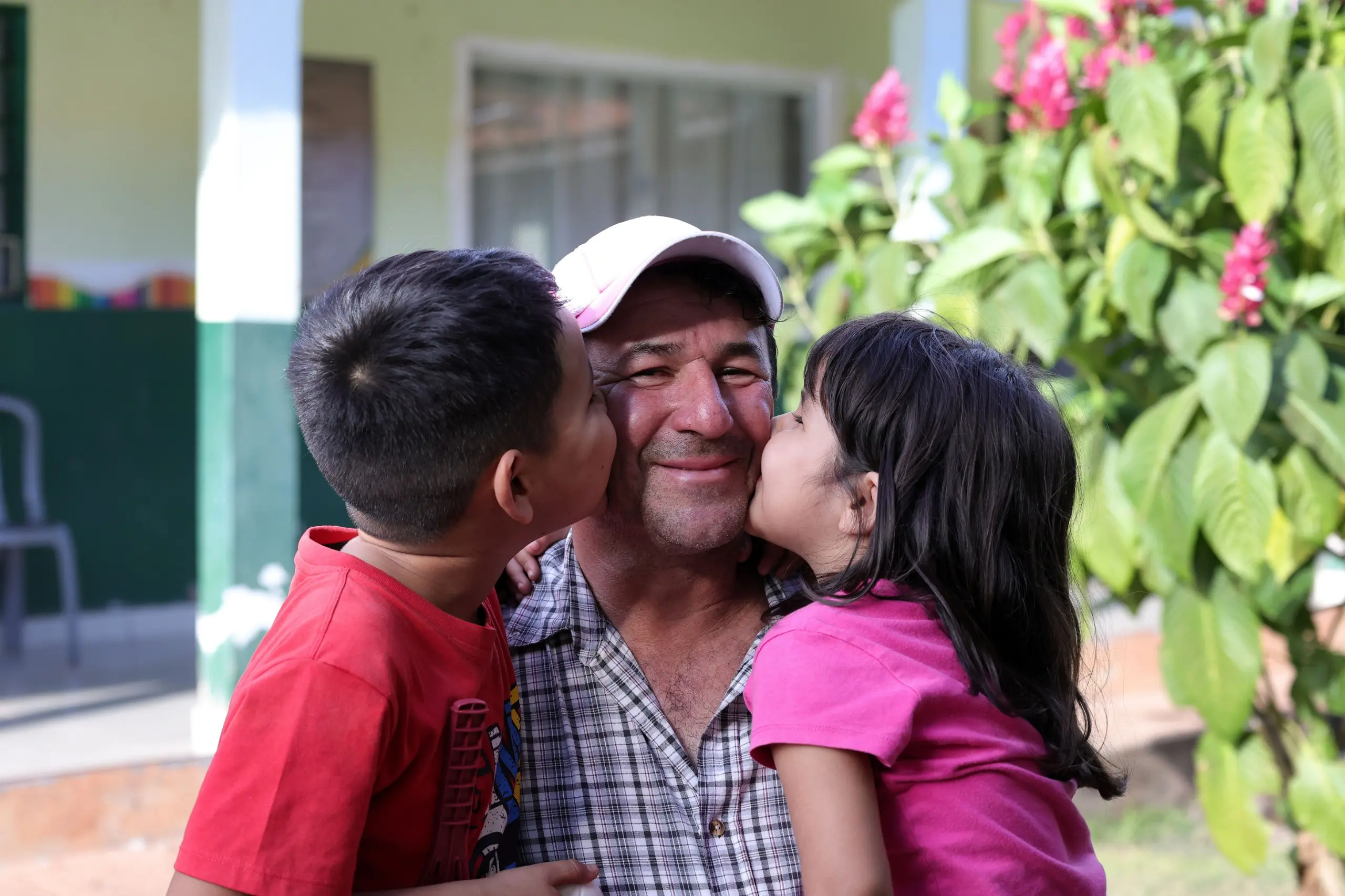 Um homem sorridente de boné claro e camisa xadrez recebe um beijo no rosto de cada lado: à esquerda, um menino de camiseta vermelha, e à direita, uma menina de camiseta rosa. Fundo externo desfocado.