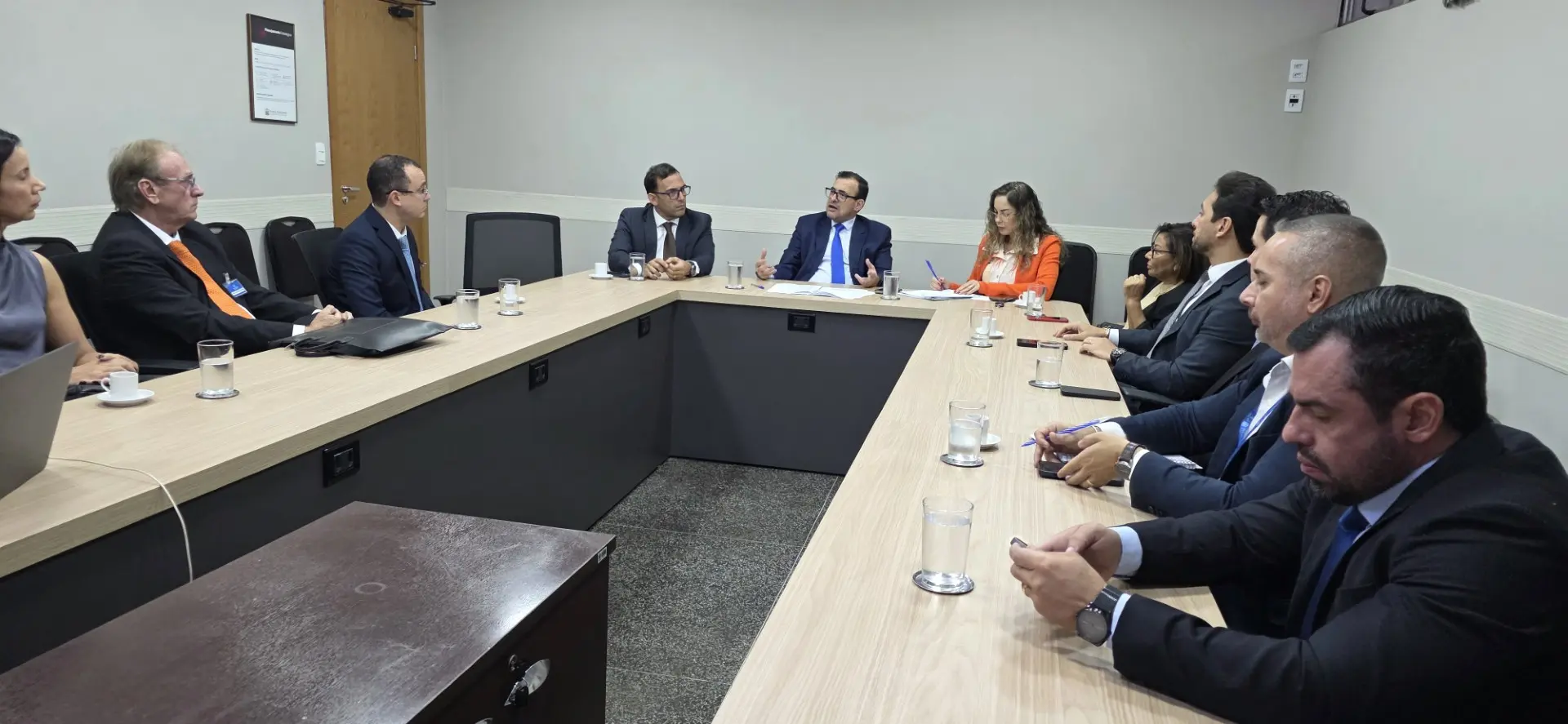 Foto horizontal que mostra os participantes de uma reunião sentados ao redor de uma mesa em formato de 