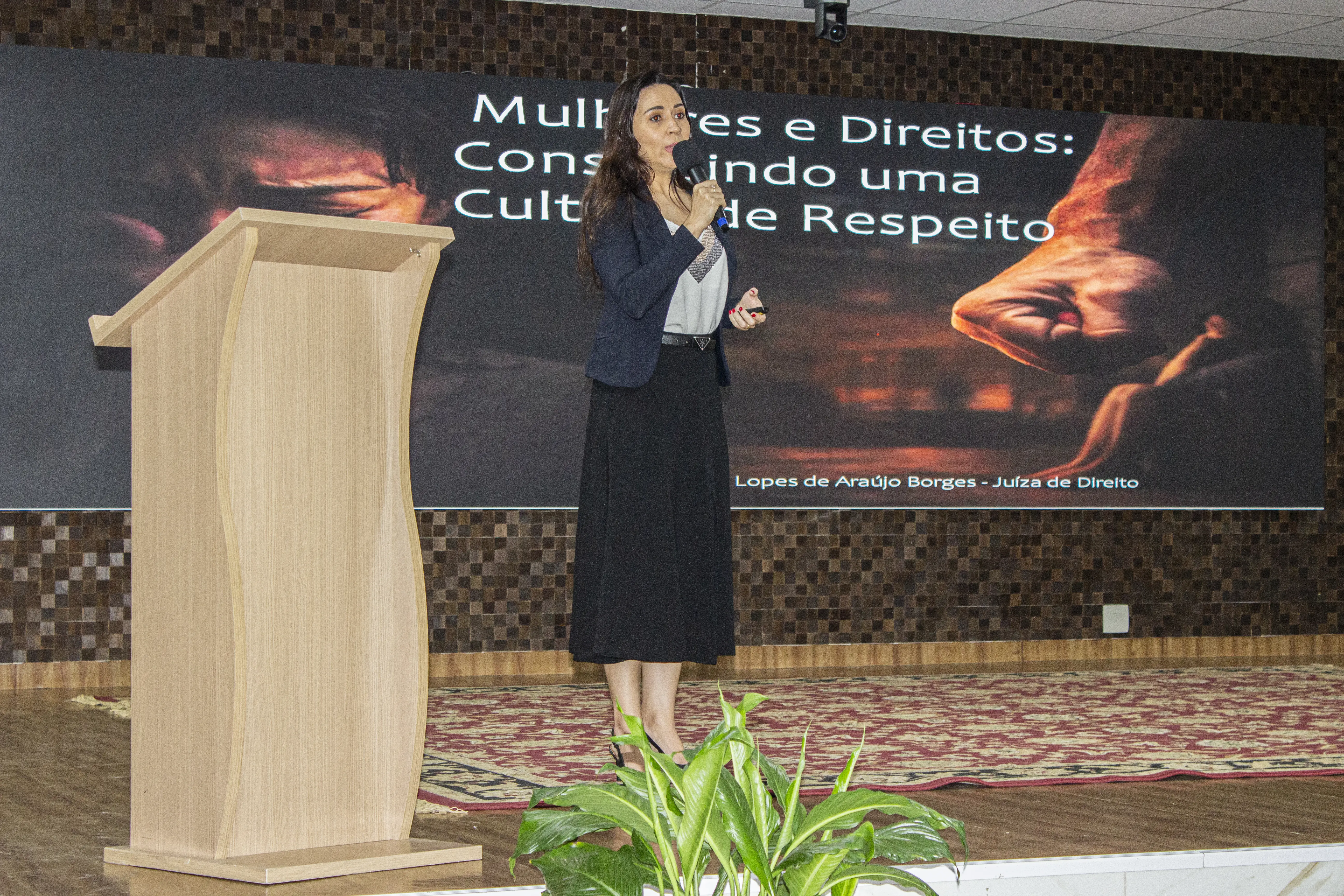 Juíza Tatyana Borges - Mulher vestindo blazer escuro, blusa clara e saia preta discursa em pé no palco com um microfone na mão. Ao lado dela, há um púlpito de madeira e, ao fundo, um grande telão com o título 