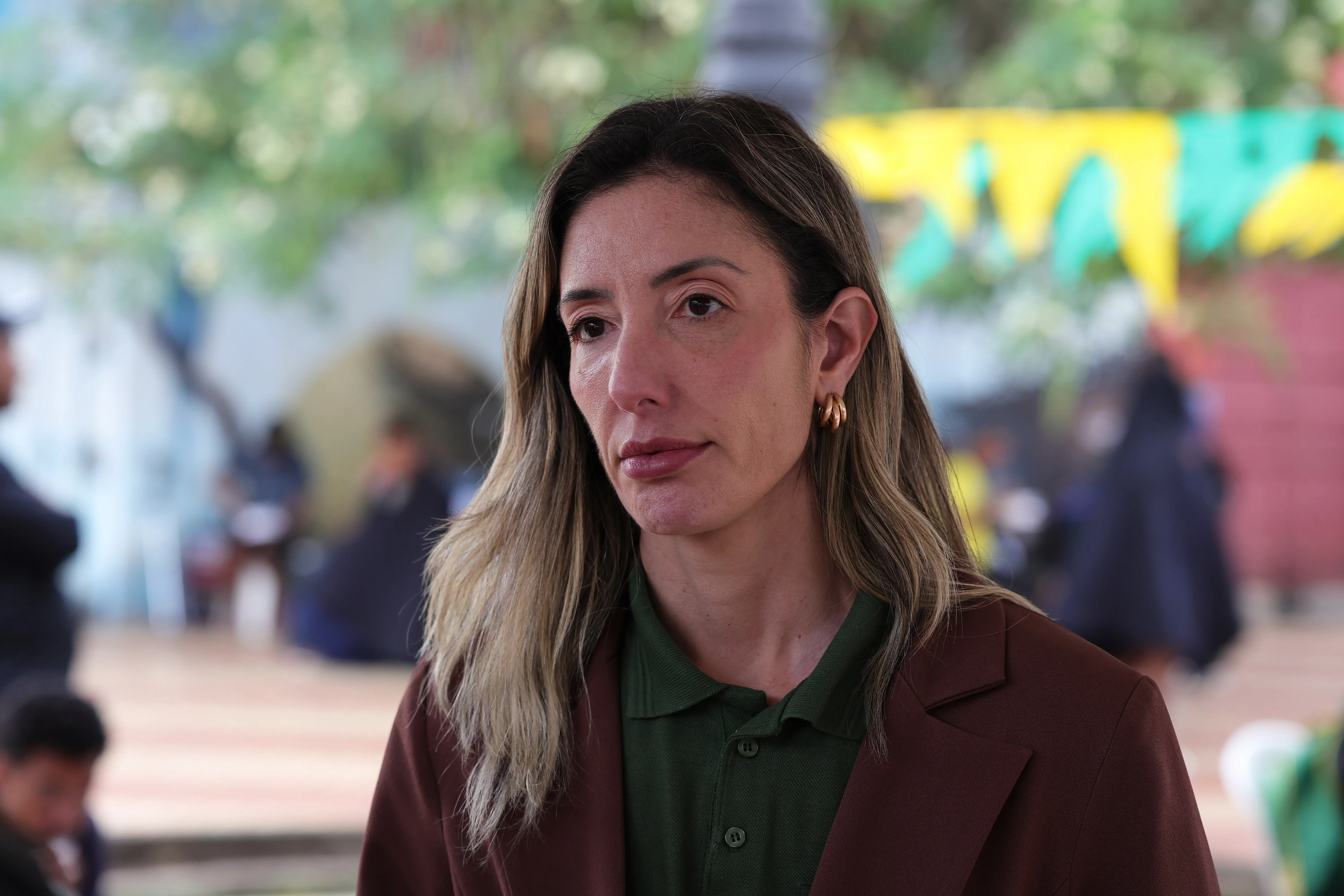Mulher com cabelos loiros e castanhos ondulados. Ela está voltada para a esquerda, com uma expressão séria. Ela veste um casaco marrom e uma blusa polo verde por baixo. O fundo está embaçado, com pessoas sentadas e vegetação, além de algumas faixas coloridas.