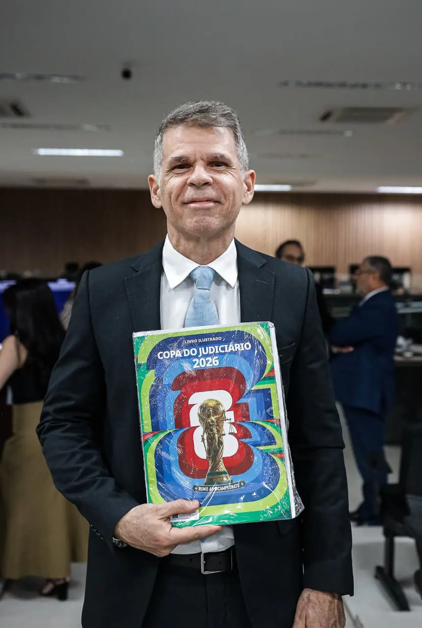 O juiz diretor do Fórum de Várzea Grande, Ângelo Judai Júnior, posa para fotografia segurando um exemplar do álbum físico da Copa do Judiciário 2026. O magistrado veste terno escuro com gravata azul clara e está em um auditório da unidade judiciária, enquanto outras pessoas aparecem ao fundo.