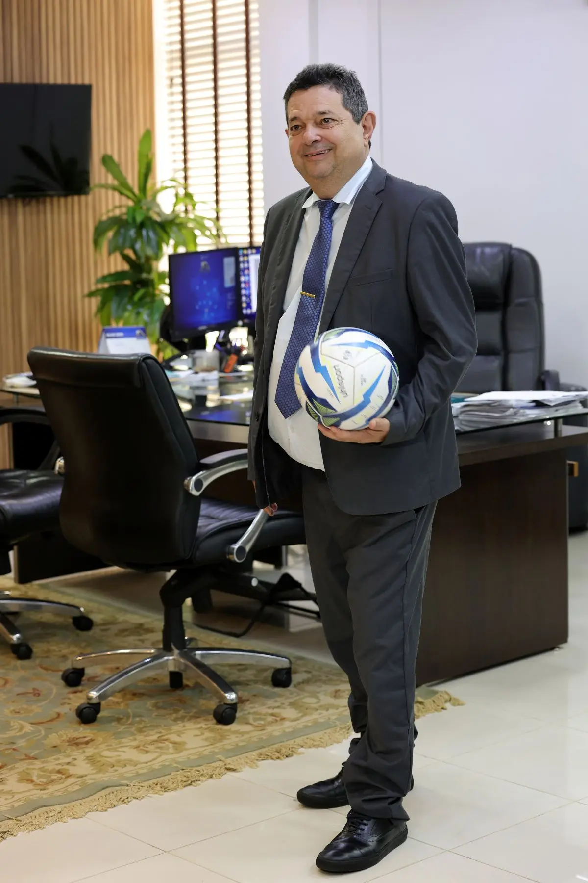 Foto vertical que mostra o desembargador José Luiz Leite Lindote em pé, sorrindo e segurando uma bola de futebol, no seu gabinete. Ele é um homem branco, de cabelo grisalho, usando terno cinza, camisa branca, gravata e sapato social preto.