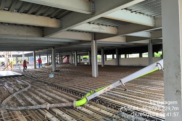 Interior de obra de edifício-garagem com estrutura metálica exposta. No chão, malha de aço para laje. Tubulação verde de bomba de concreto ao centro. Operários ao fundo. Carimbo de data: 26/06/2026.