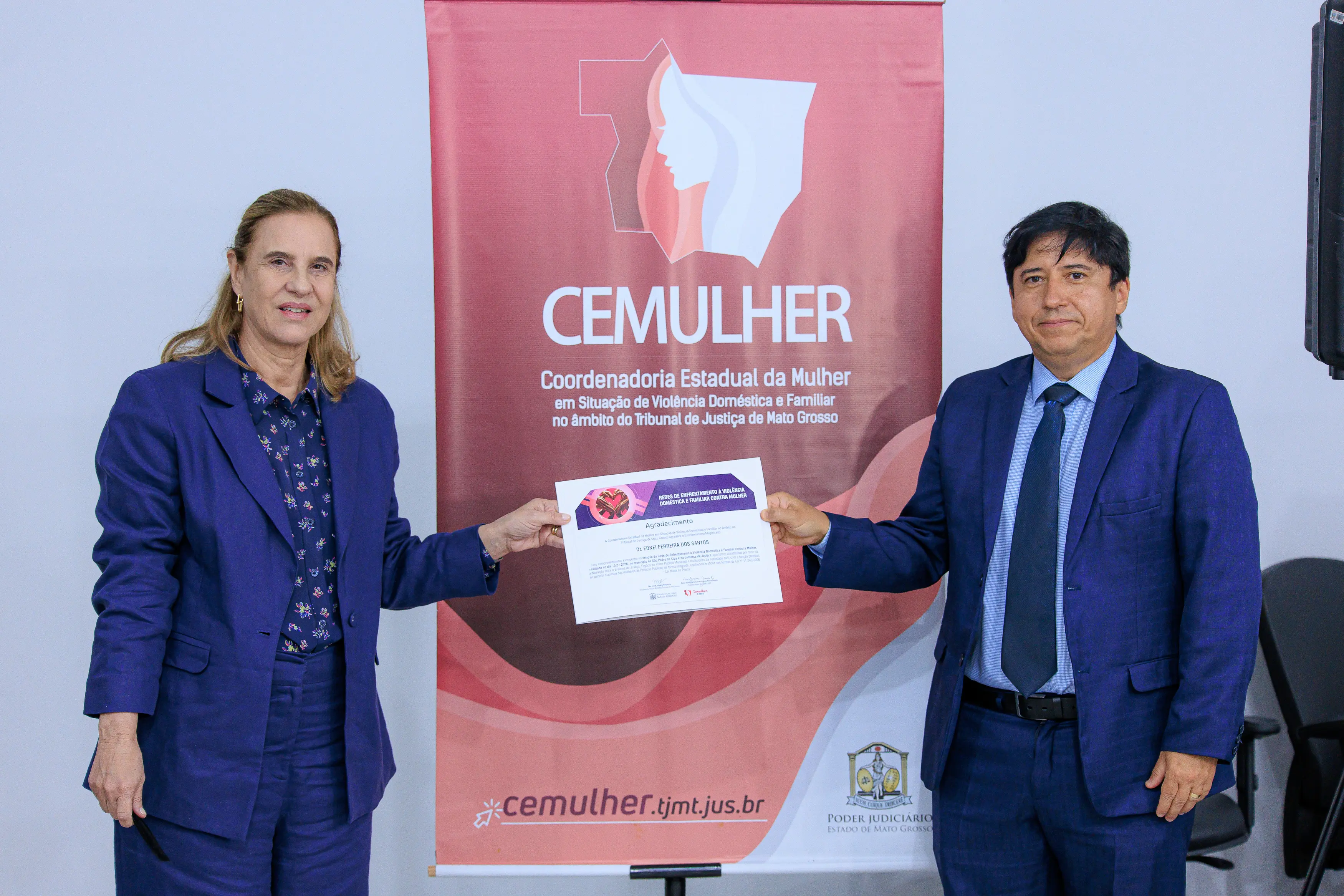 Desembargadora Vandymara Galvão e juiz Edinei Ferreira estão de pé lado a lado, segurando juntos um certificado. Ambos vestem ternos azuis. Atrás deles, há um banner de pé vermelho e branco com o texto 